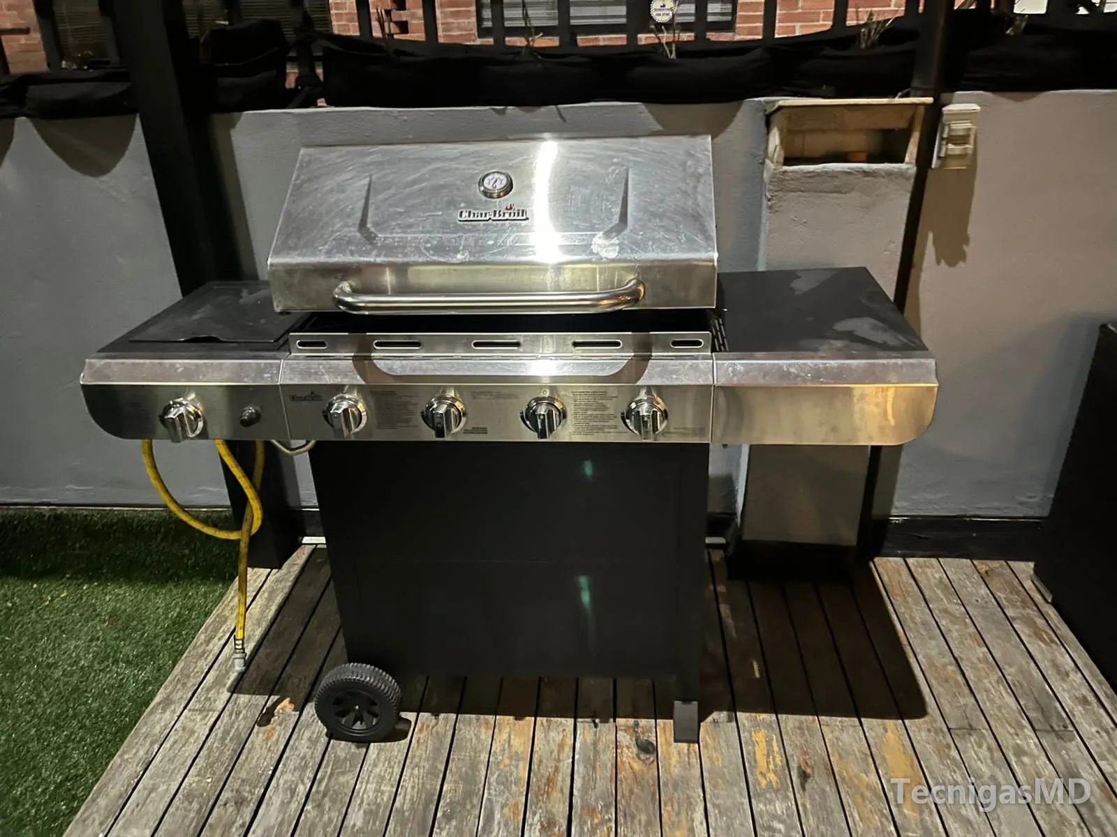 Asador de acero inoxidable de cuatro quemadores y hornilla lateral sobre deck de madera, con manguera amarilla de gas