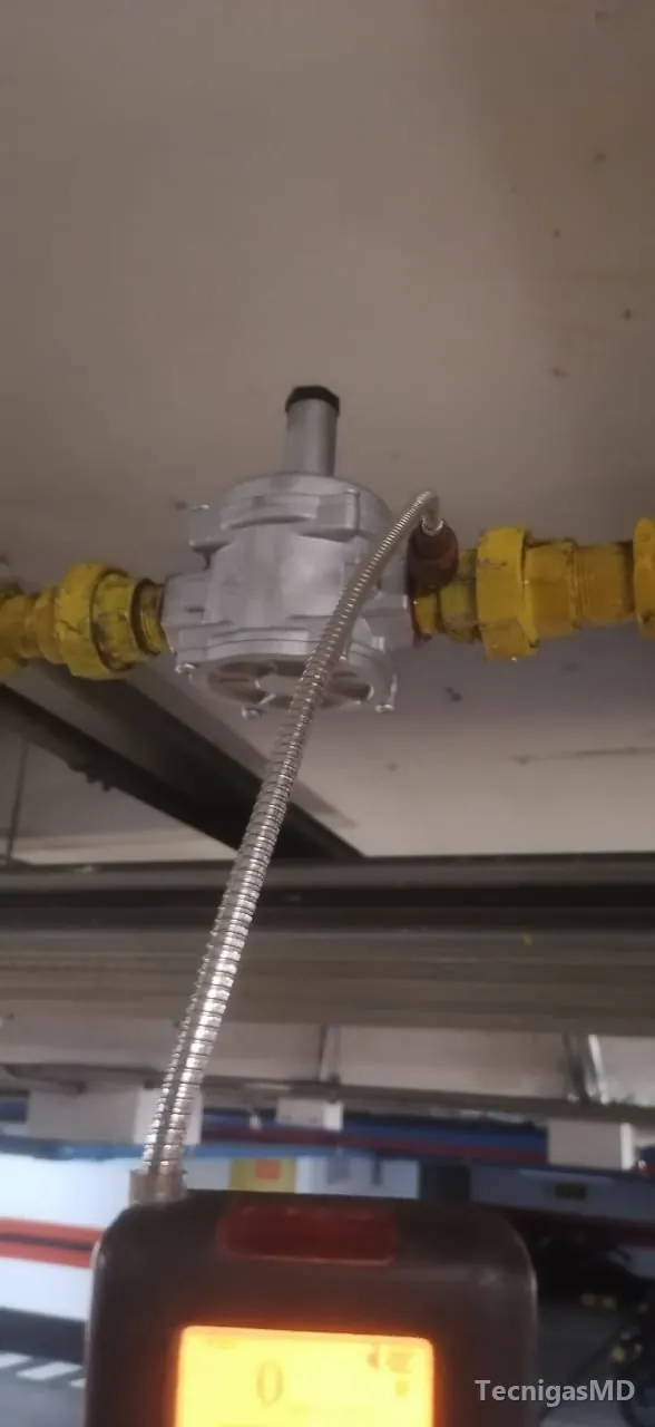 Detector electrónico con sonda flexible midiendo junto a un regulador en tubería amarilla bajo el techo de un parqueadero