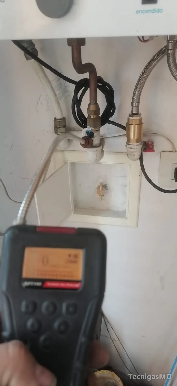 Explosímetro con sonda flexible revisando las conexiones de cobre y mangueras metálicas bajo un calentador a gas blanco