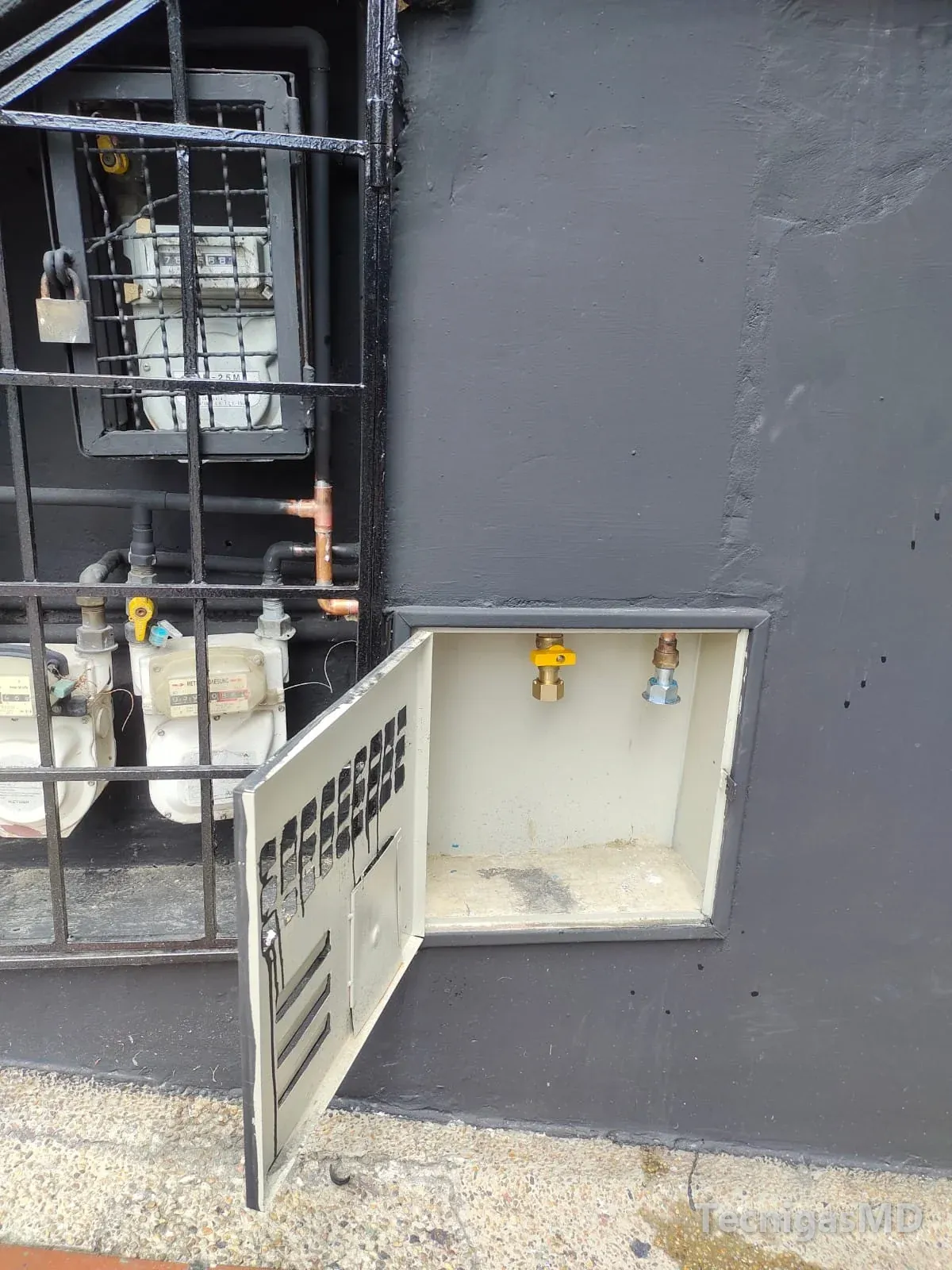 Gabinete abierto en fachada gris oscuro con válvula de corte y acople listos para el medidor, junto a medidores tras reja