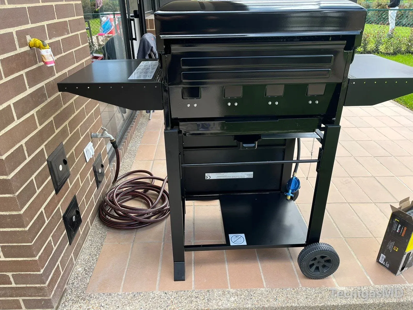 Asador a gas visto por detrás en una terraza, junto a un muro de ladrillo con la salida del punto de gas envuelta en cinta