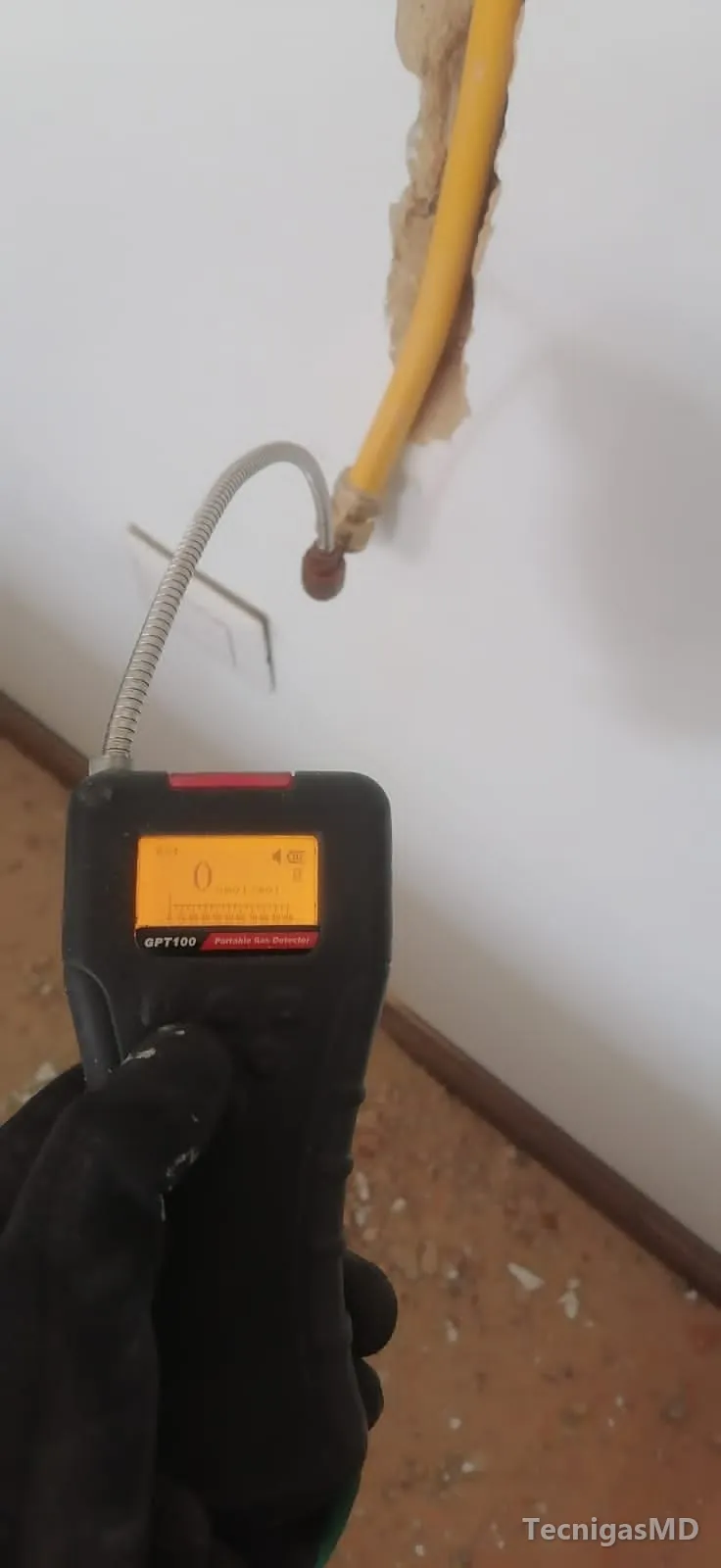 Mano con guante sosteniendo un detector de gas en el conector de bronce de una tubería amarilla que sale de un muro en obra
