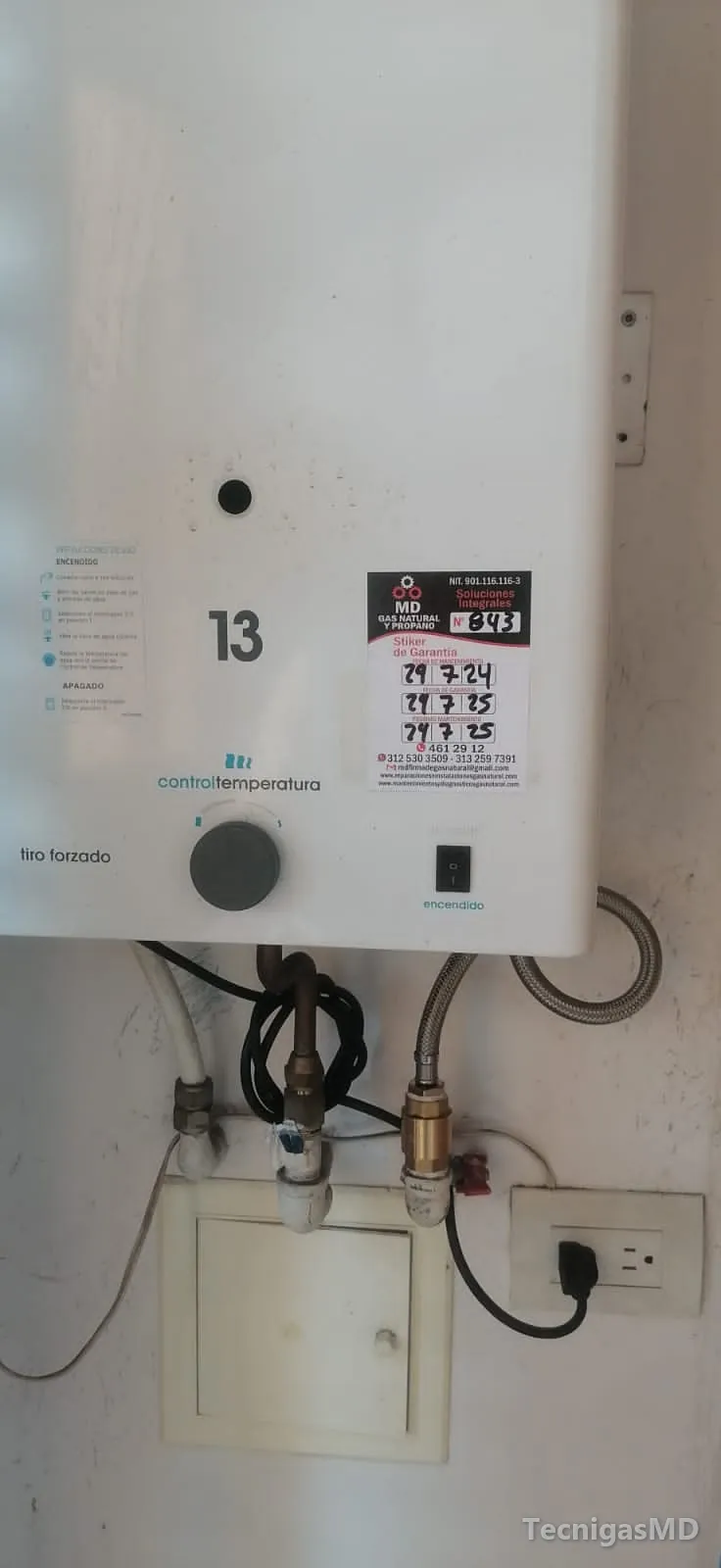 Parte inferior de un calentador a gas de tiro forzado con adhesivo de garantía, conexiones de agua y gas y tomacorriente