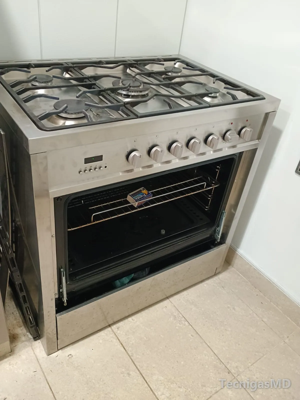Estufa de piso a gas en acero inoxidable con cinco quemadores, perillas frontales y horno abierto con su parrilla