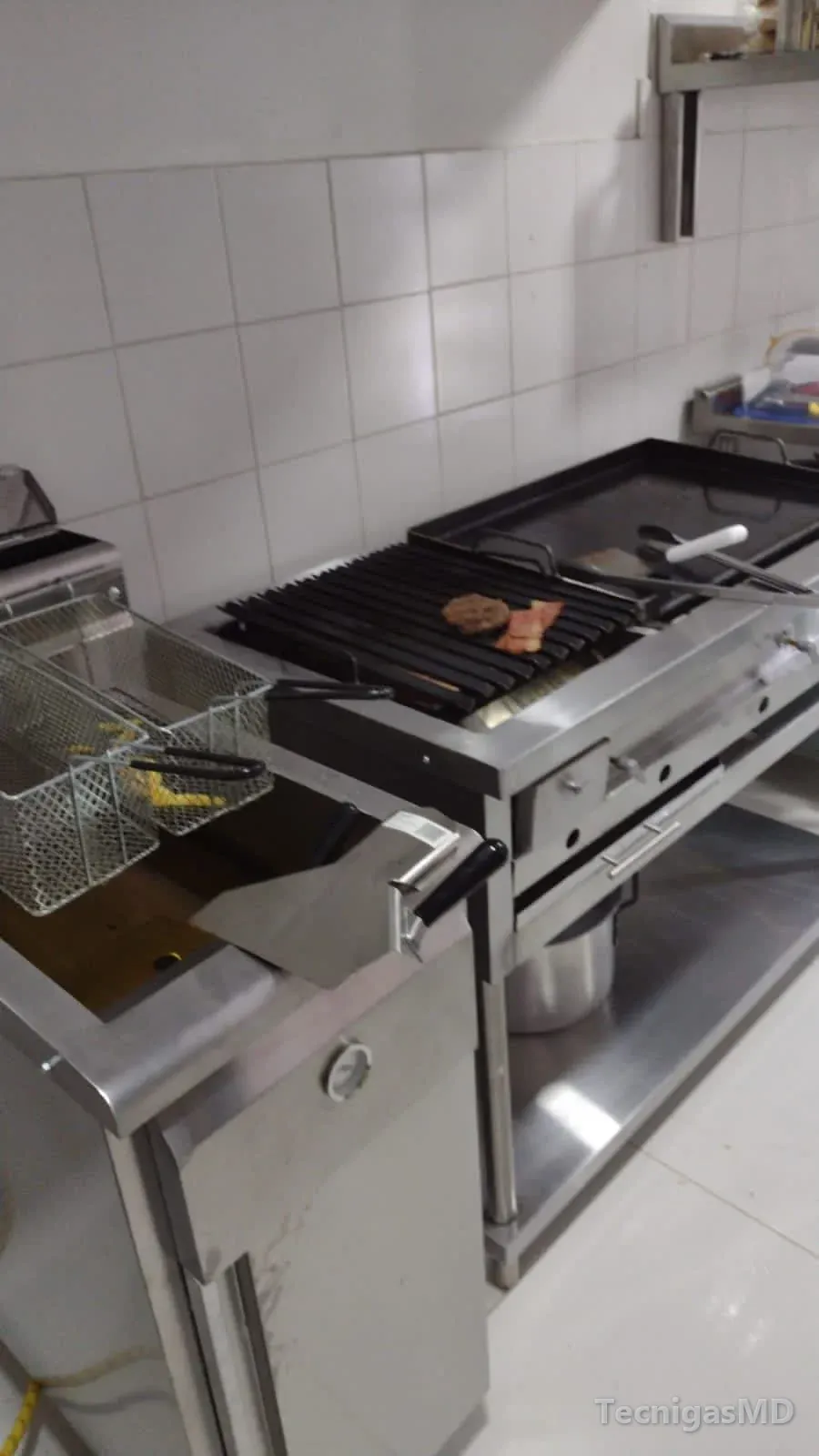 Línea de cocina comercial con freidora de canastillas, parrilla asadora con carne y plancha a gas en acero inoxidable