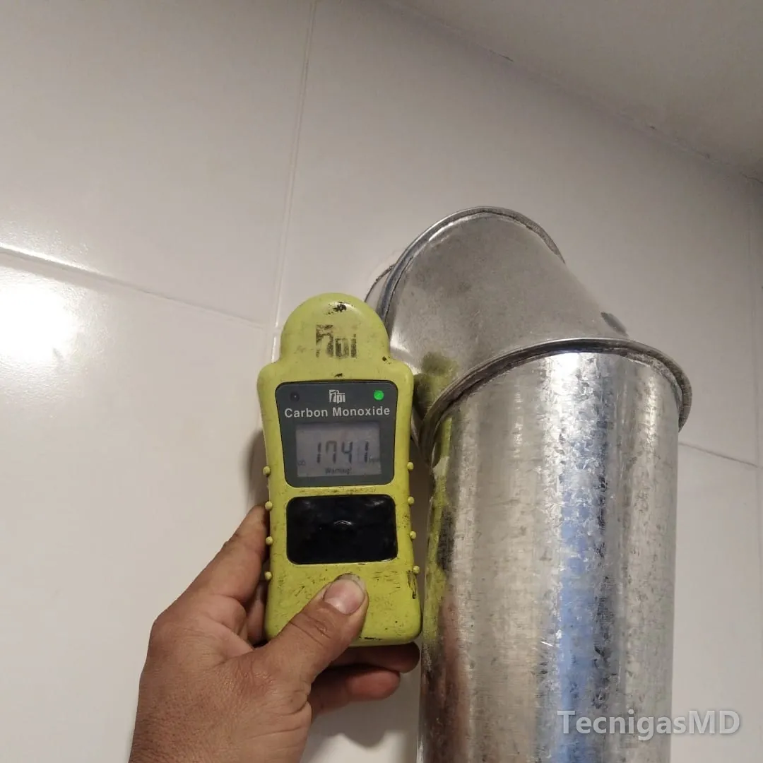 Mano con detector de monóxido de carbono junto al codo de un ducto galvanizado de evacuación de gases, cerca del techo