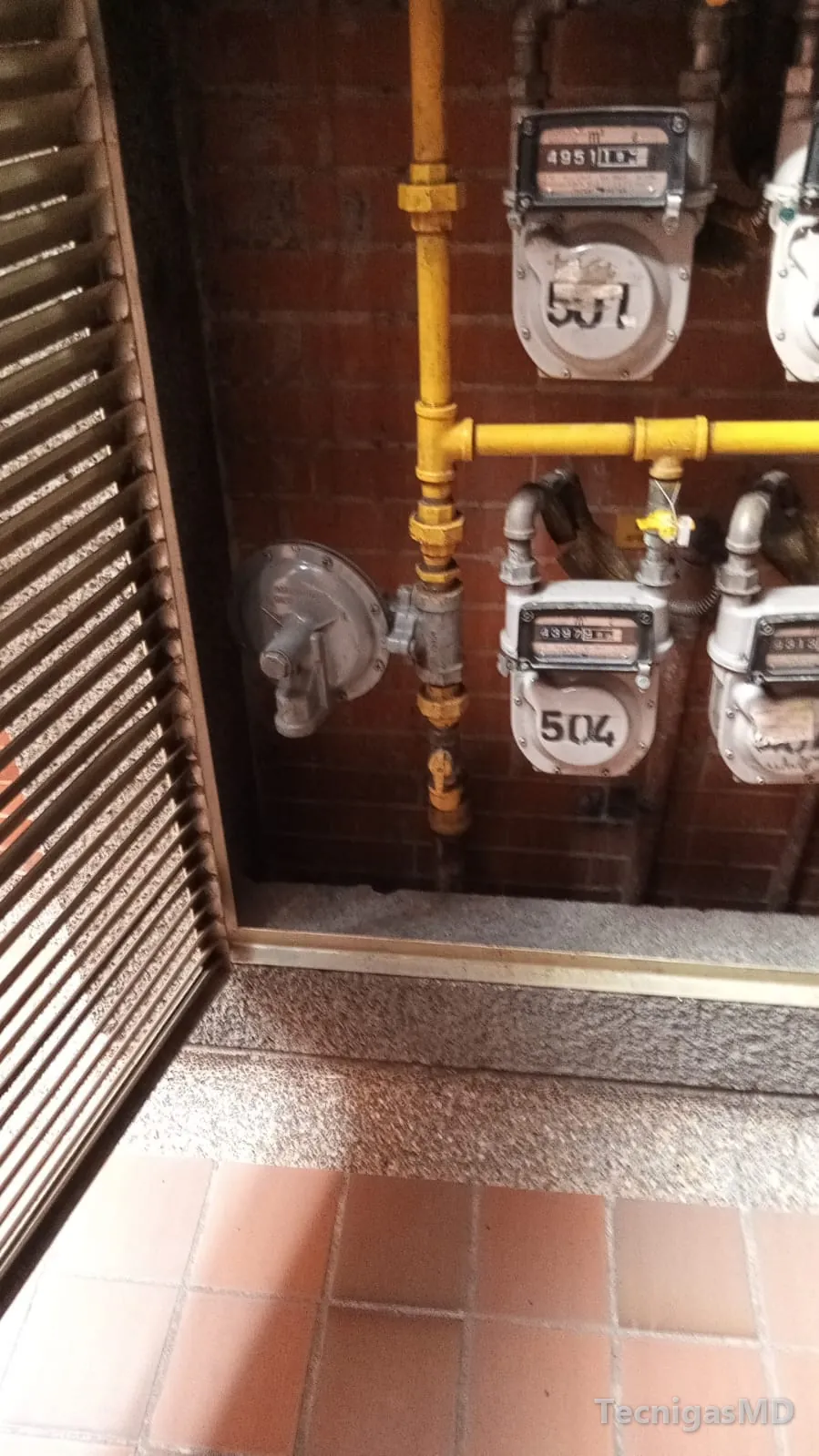 Medidores de gas natural numerados con regulador y tubería amarilla en un centro de medición con puerta de rejilla