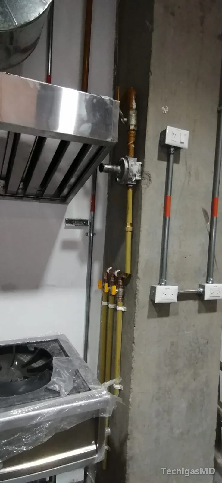 Tubería amarilla con regulador y tres salidas con válvula, junto a una estufa industrial aún con plástico y su campana
