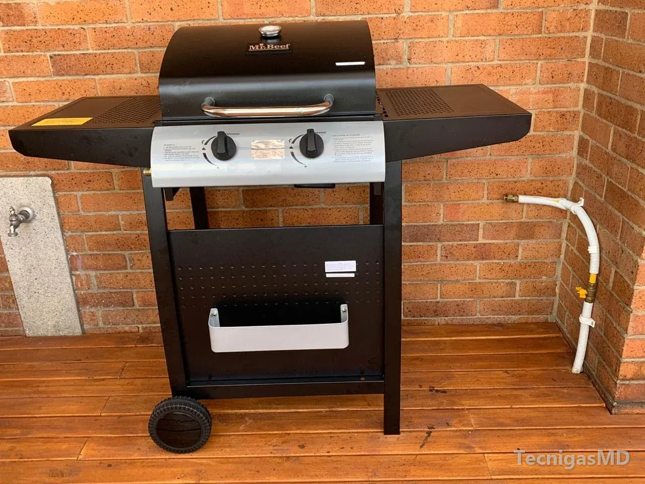 Asador a gas de dos quemadores en deck de madera junto al punto de gas: tubería flexible blanca, válvula amarilla y acople