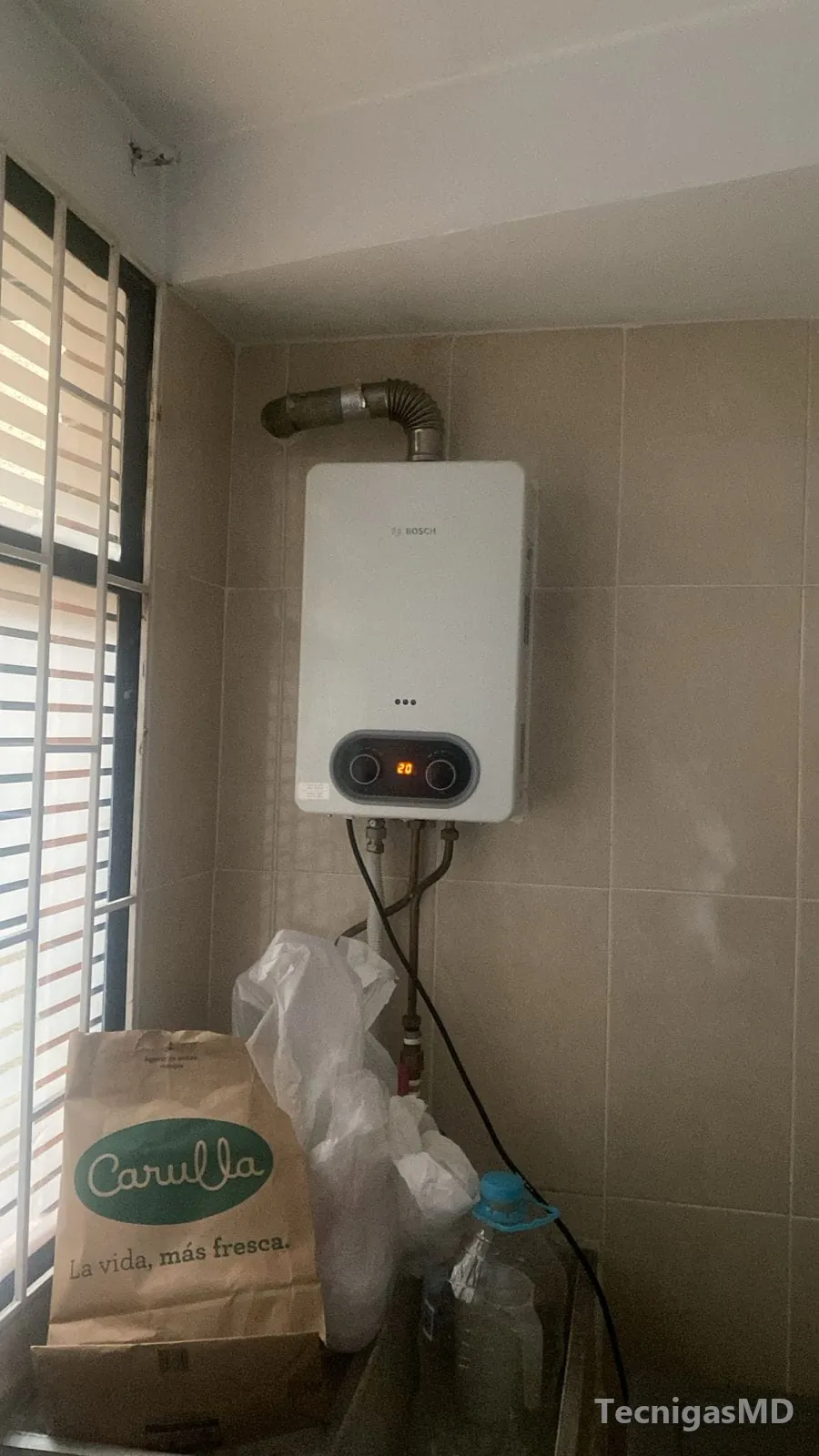 Calentador a gas Bosch con pantalla digital y ducto de evacuación metálico, instalado en muro enchapado junto a una ventana