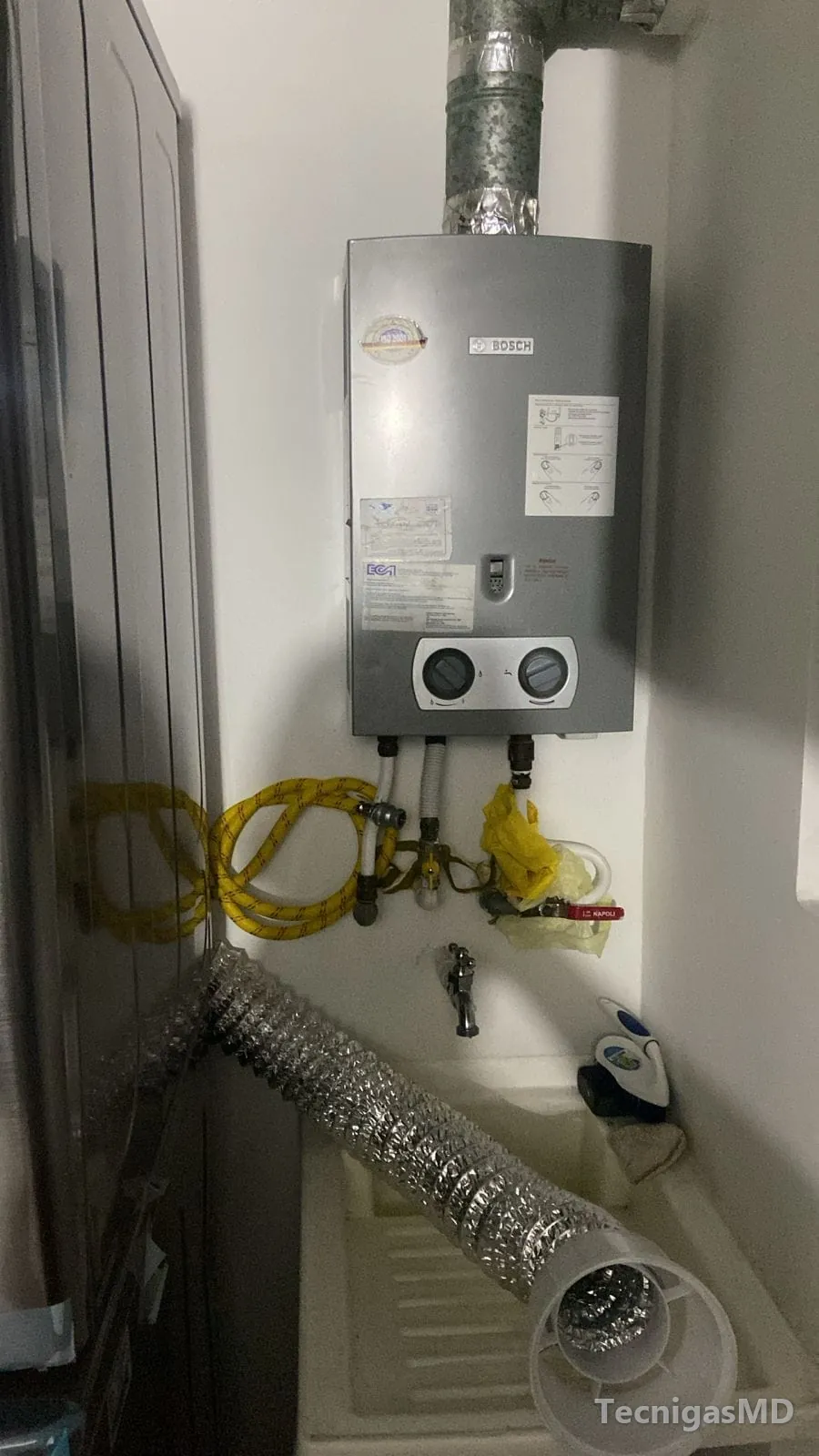 Calentador a gas de paso Bosch junto a un lavadero, con ducto de aluminio, mangueras flexibles amarillas y válvulas