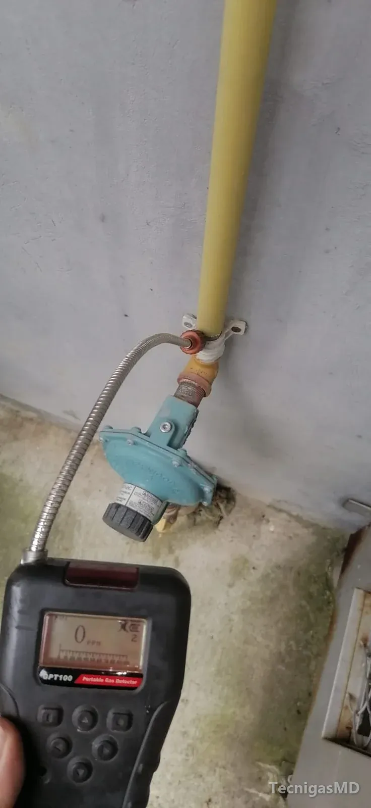 Detector portátil de gas con sonda flexible junto a un regulador azul en la base de una tubería amarilla de gas natural