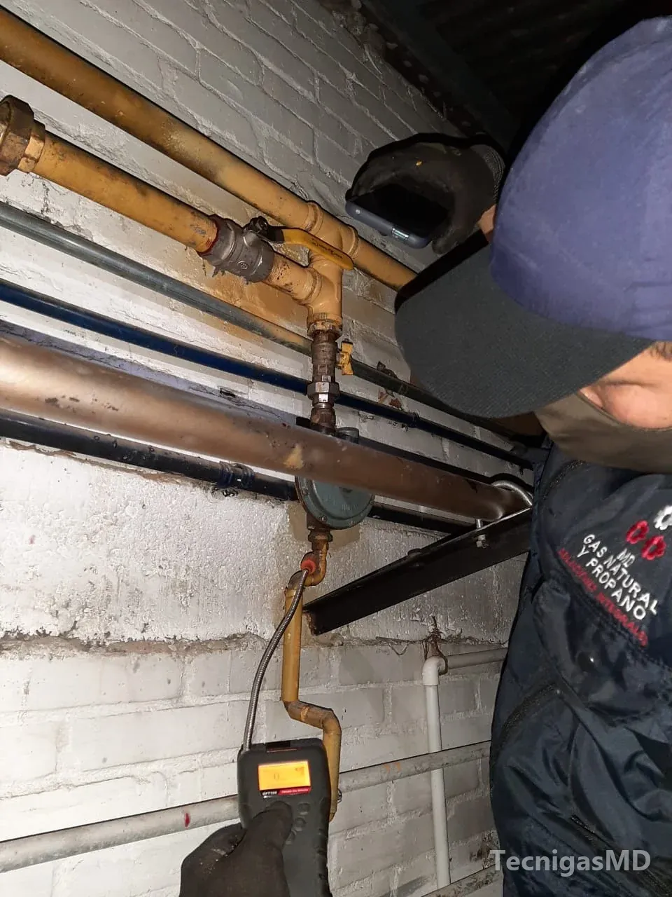 Técnico con guantes que sostiene un celular y acerca un detector electrónico de gas a un regulador en tubería amarilla