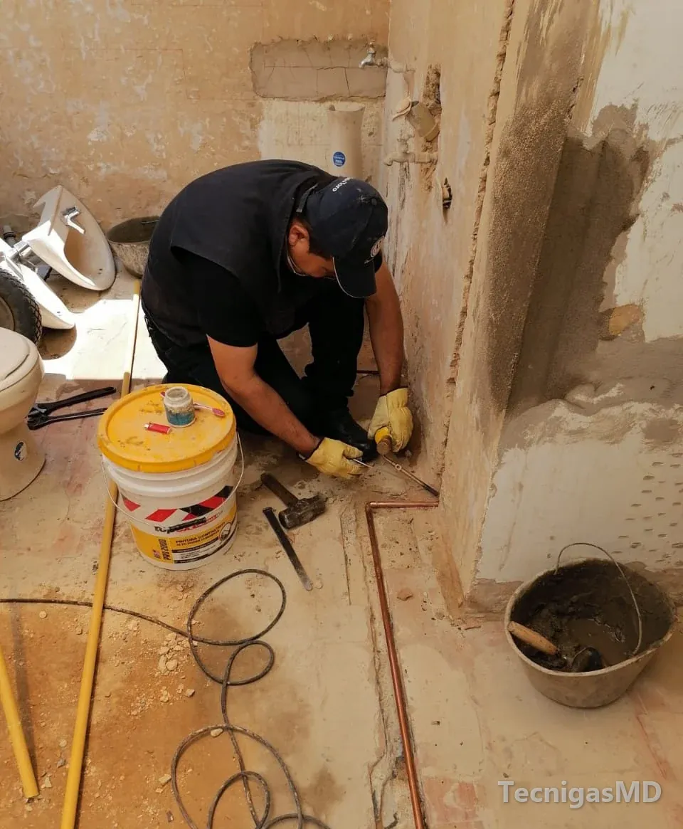 Técnico con guantes trabajando al pie del muro sobre tubería de cobre en un baño en remodelación, con sanitarios desmontados